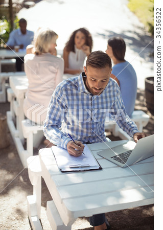 Man writing on clipboard while using laptop Man writing on clipboard while using laptop 34356522