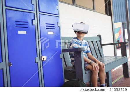 Boy using virtual reality glasses while sitting on bench by lockers 34358729