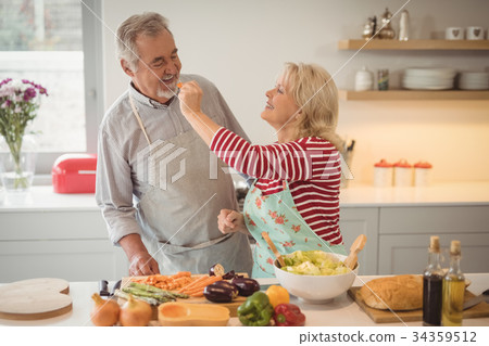 Senior woman feeding vegetable salad to man in kitchen 34359512