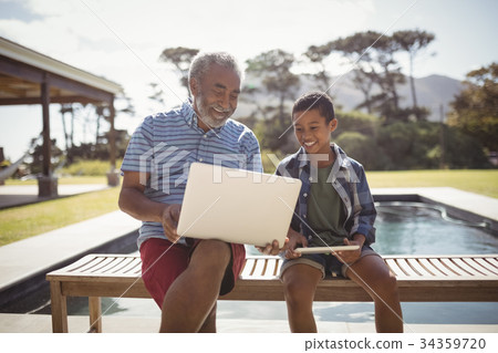 Smiling grandfather showing laptop to grandson near poolside 34359720