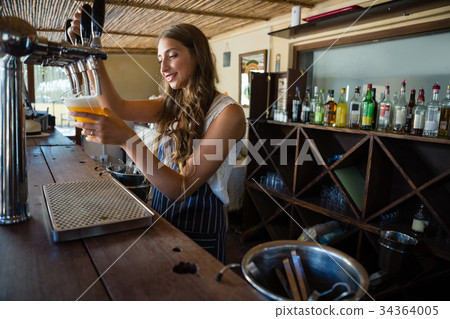 Barmaid pouring beer from tap in glass at bar 34364005