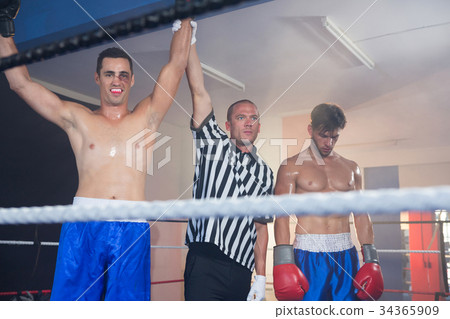 Referee holding hands of smiling male boxing winner by athlete 34365909