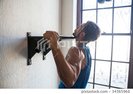 Side view of male athlete practicing pull ups on bar by window Side view of male athlete practicing pull ups on bar by window 34365968