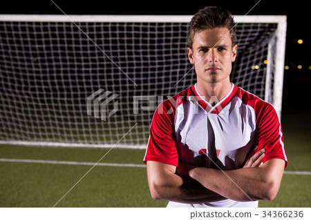 Confident male soccer player standing with arms crossed against goal post on field 34366236