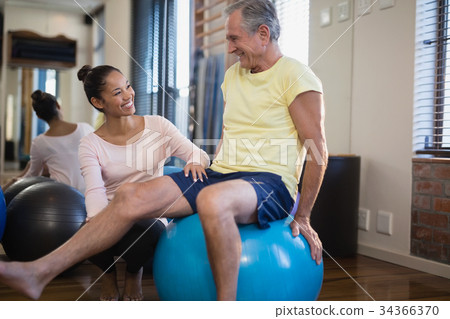 Smiling female therapist crouching by senior male patient sitting on exercise ball 34366370