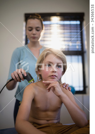 Thoughtful shirtless boy sitting with female therapist using ultrasound machine on shoulder Thoughtful shirtless boy sitting with female therapist using ultrasound machine on shoulder 34366538