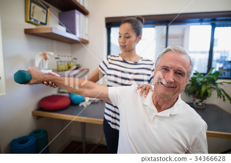 Female doctor holding hand of smiling senior male patient lifting dumbbell Female doctor holding hand of smiling senior male patient lifting dumbbell 34366628