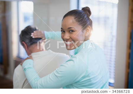 Rear view of portrait of smiling female therapist giving neck massaging to senior male patient 34366728