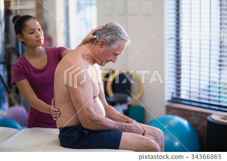 Young female therapist giving back massage to senior male patient sitting on bed Young female therapist giving back massage to senior male patient sitting on bed 34366865