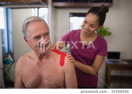 Female therapist applying elastic therapeutic tape on shoulder of smiling shirtless senior male pati 34366886