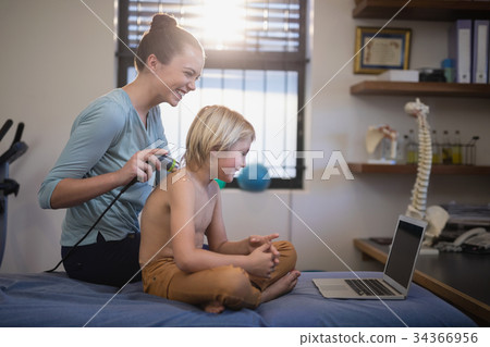 Cheerful female therapist while scanning shoulder of shirtless boy looking at laptop 34366956