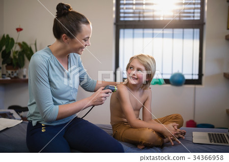 Smiling young female therapist scanning shoulder while looking at shirtless boy sitting on bed 34366958