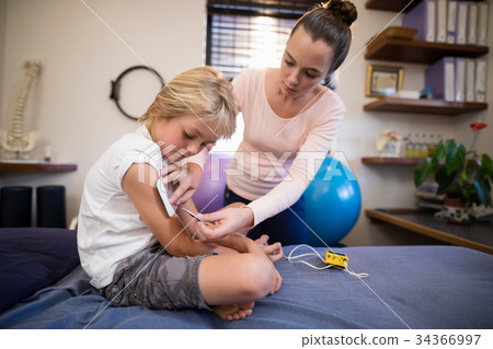 Young female therapist positioning electrodes on arm of boy Young female therapist positioning electrodes on arm of boy 34366997