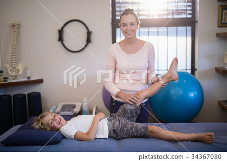 Portrait of smiling young female therapist examining knee with boy lying on bed Portrait of smiling young female therapist examining knee with boy lying on bed 34367080