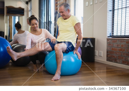 Female therapist helping senior male patient doing leg exercise on blue ball 34367100