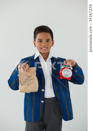 Schoolboy holding alarm clock and disposable lunch bag against white background Schoolboy holding alarm clock and disposable lunch bag against white background 34367239