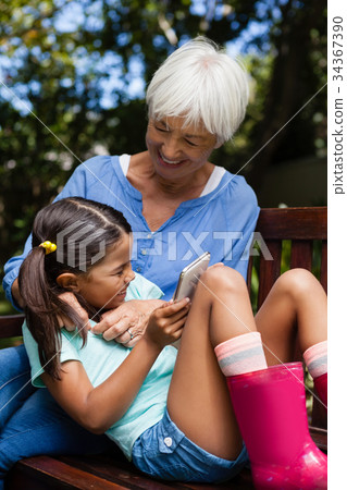 Cheerful senior woman enjoying with girl holding mobile phone on bench Cheerful senior woman enjoying with girl holding mobile phone on bench 34367390