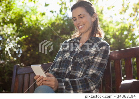 Low angle view of smiling beautiful woman text messaging while sitting on wooden bench 34368194
