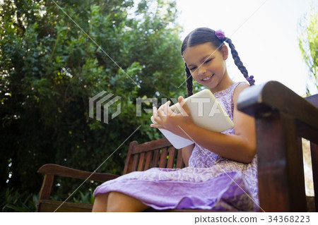 Low angle view of smiling girl using digital tablet while sitting on wooden bench 34368223