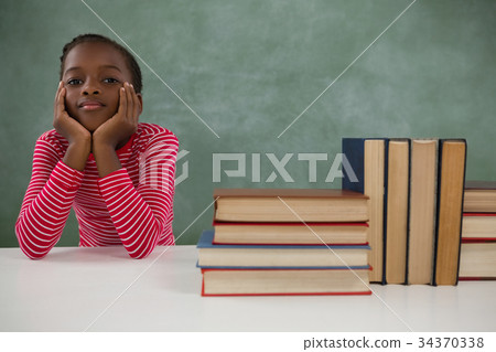 Schoolgirl sitting beside books stack against chalkboard 34370338