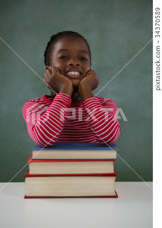 Schoolgirl leaning on books stack against chalkboard in classroom 34370589