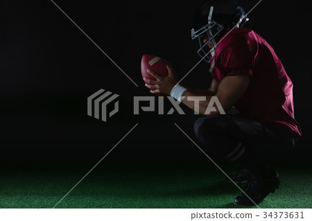 American football player sitting on toes holding a ball with both his hands 34373631