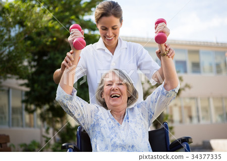 Doctor assisting woman in lifting dumbbells at park Doctor assisting woman in lifting dumbbells at park 34377335