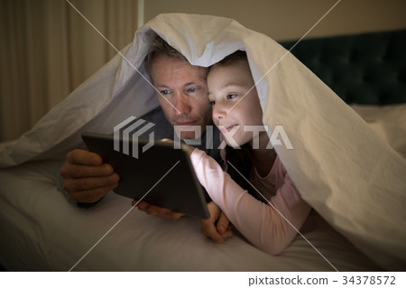 Father and daughter using digital tablet under blanket on bed Father and daughter using digital tablet under blanket on bed 34378572