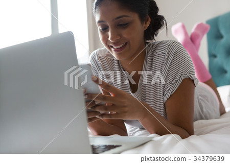 Smiling woman with laptop using phone on bed Smiling woman with laptop using phone on bed 34379639