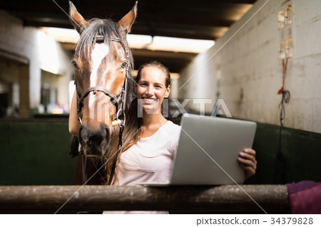 Portrait of female jockey holding laptop while standing by horse 34379828