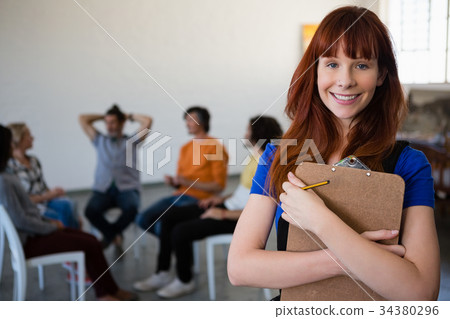 Portrait of happy teacher holding clipboard with students talking in background 34380296