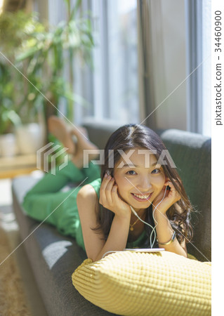 A woman with long hair who enjoys the music downloaded by a smartphone lying on a sofa by the window with a pleasant sun 34460900