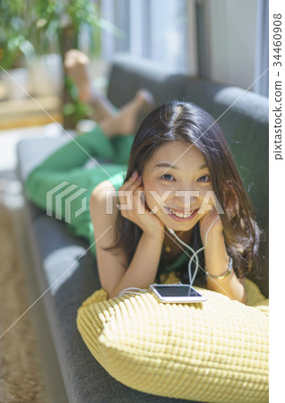 一個長頭髮的女人喜歡用智能手機下載的音樂，在陽光舒適的窗戶旁躺在沙發上 34460908