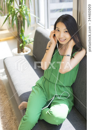 一個長頭髮的女人喜歡用智能手機下載的音樂，在陽光舒適的窗戶旁躺在沙發上 34460909