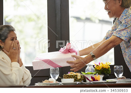 a senior man is making surprise to his wife in the restaurant by giving a gift 34530933