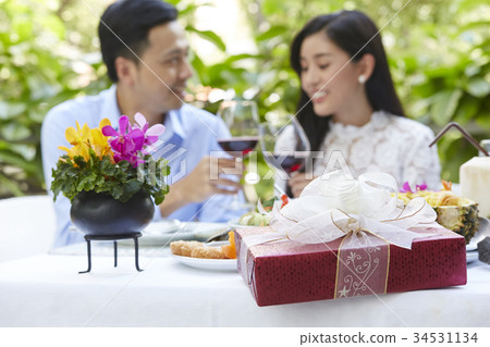 Two people are sitting on a table with food and gift and holding glasses of wine 34531134