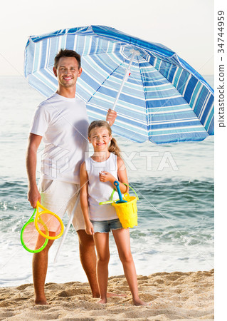 Young dad with girl standing on beach with toys Young dad with girl standing on beach with toys 34744959