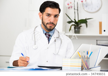 Young man doctor is working behind laptop and reading documents Young man doctor is working behind laptop and reading documents 34749831