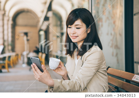 Woman watching smartphone while drinking coffee on a bench Woman watching smartphone while drinking coffee on a bench 34843013