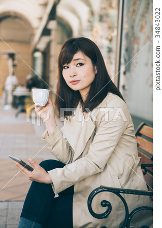 Woman watching smartphone while drinking coffee on a bench Woman watching smartphone while drinking coffee on a bench 34843022