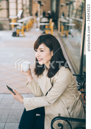 婦女觀看的智能手機,當喝在長凳時的咖啡 婦女觀看的智能手機,當喝在長凳時的咖啡 34843023