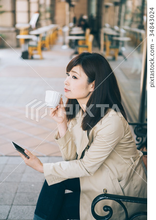 婦女觀看的智能手機,當喝在長凳時的咖啡 婦女觀看的智能手機,當喝在長凳時的咖啡 34843024