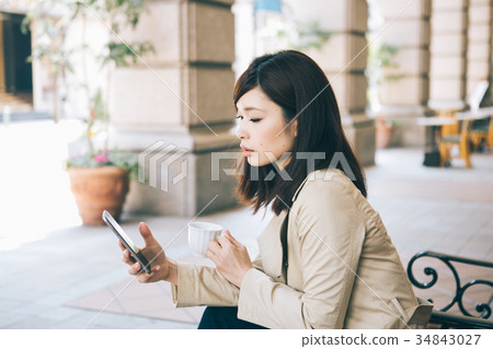 Woman watching smartphone while drinking coffee on a bench Woman watching smartphone while drinking coffee on a bench 34843027