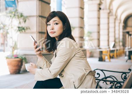 婦女觀看的智能手機,當喝在長凳時的咖啡 婦女觀看的智能手機,當喝在長凳時的咖啡 34843028
