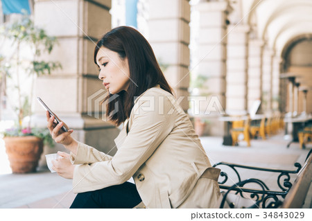 Woman watching smartphone while drinking coffee on a bench Woman watching smartphone while drinking coffee on a bench 34843029