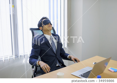 Young sales businessman in suit wearing eye mask and taking a nap break at company office Young sales businessman in suit wearing eye mask and taking a nap break at company office 34959866