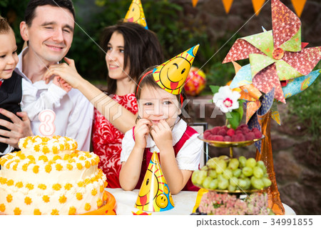 Children's birthday party girl sitting at a table near a cake in a festive hat and smiling Children's birthday party girl sitting at a table near a cake in a festive hat and smiling 34991855