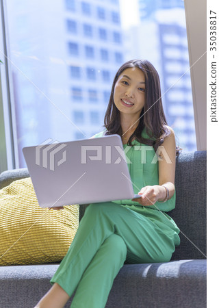 有長的頭髮的美麗的婦女坐由與宜人的陽光的窗口的沙發和與便攜式計算機一起使用 有長的頭髮的美麗的婦女坐由與宜人的陽光的窗口的沙發和與便攜式計算機一起使用 35038817