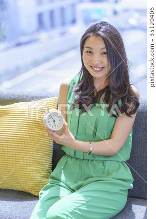 有長的頭髮的美麗的婦女坐沙發由窗口和顯示她鬧鐘教她的時間 有長的頭髮的美麗的婦女坐沙發由窗口和顯示她鬧鐘教她的時間 35120406