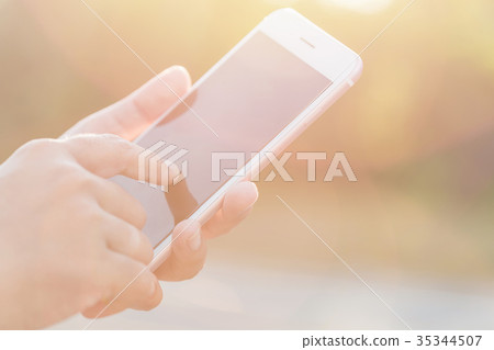 Hand of woman holding or using smartphone on green blur and sunlight effect 35344507